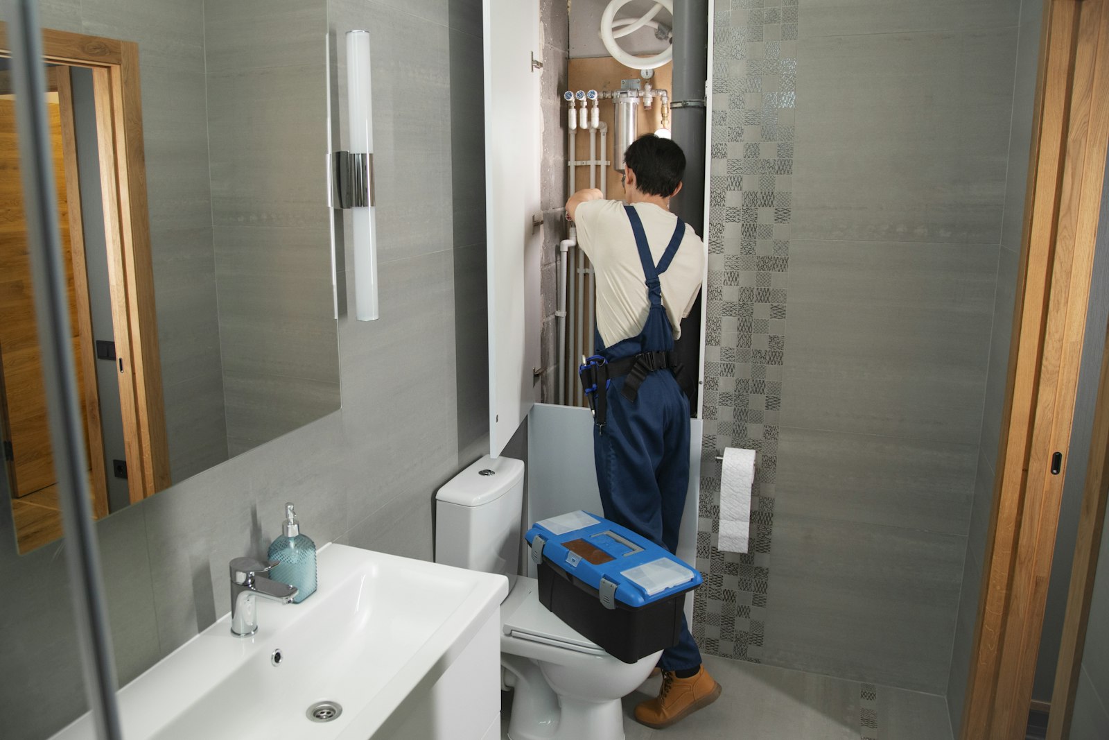 Plumber in work overalls inspecting bathroom plumbing with his toolbox beside the toilet